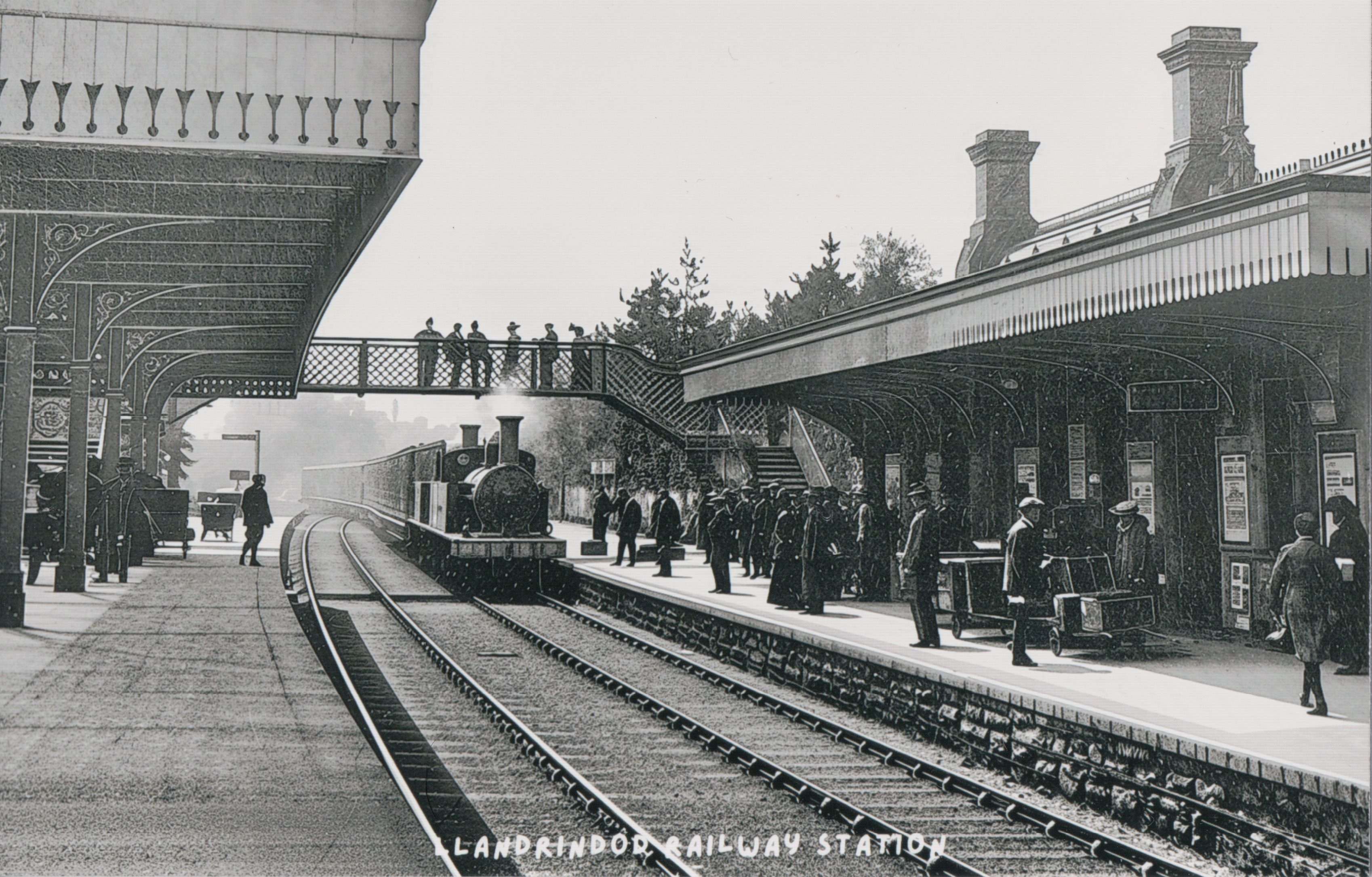 Historic view of a crowded platform at Llandrindod Wells railway station with steam locomotive, canopies and footbridge