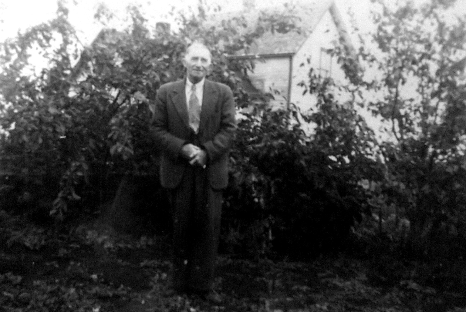 Owen Jones Davies (1875–1958), Welsh-born Canadian farmer, standing in a garden in a suit, with shrubs and a house behind him.